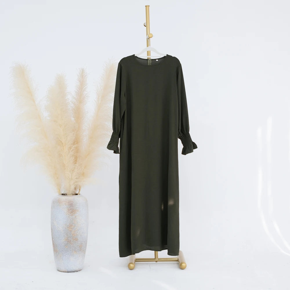 Vestido abaya holgado verde oliva oscuro colgado en percha de madera junto a un jarrón de pampas seco.