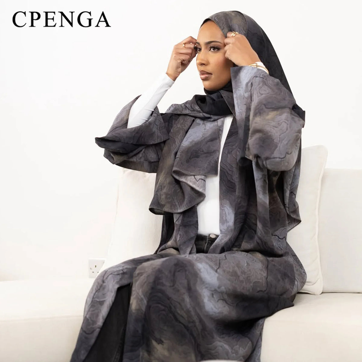 Eine Frau in einem eleganten grauen Blumen-Abaya und Hijab-Set sitzt auf einem weißen Sofa.