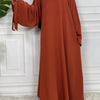 Eine Frau in einem fließenden rostfarbenen Maxi-Abaya mit langen Ärmeln vor einer weißen Wand.