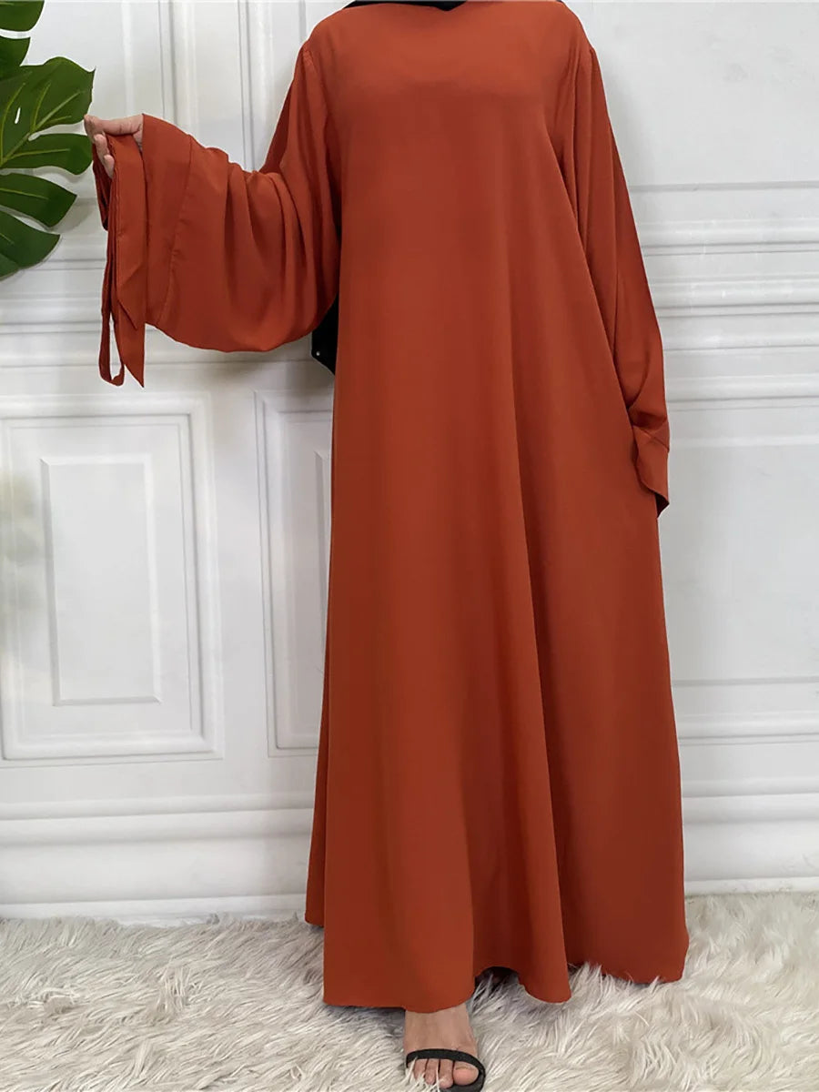 Eine Frau in einem fließenden rostfarbenen Maxi-Abaya mit langen Ärmeln vor einer weißen Wand.