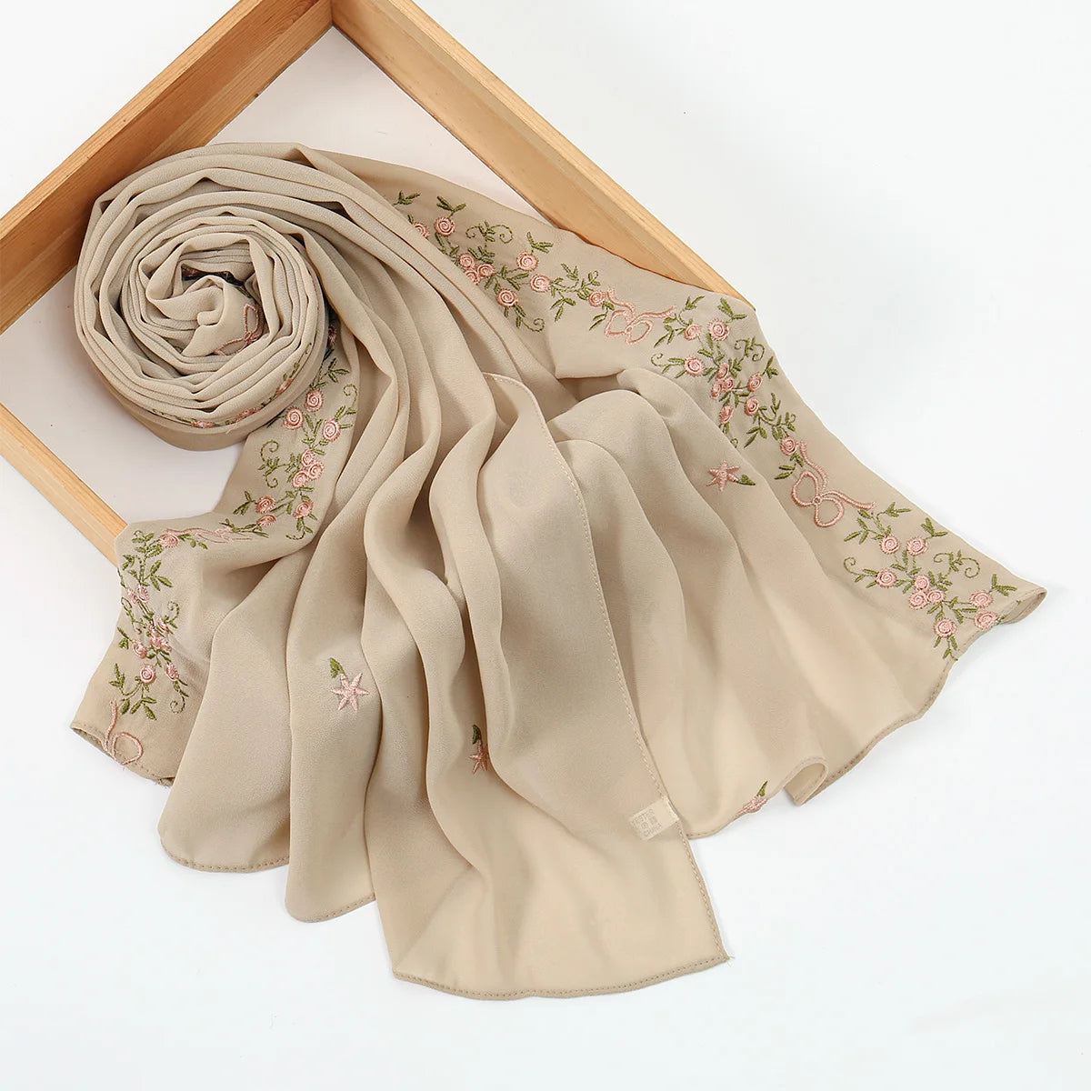 A beige chiffon hijab shawl with delicate pink floral embroidery draped over a wooden frame.