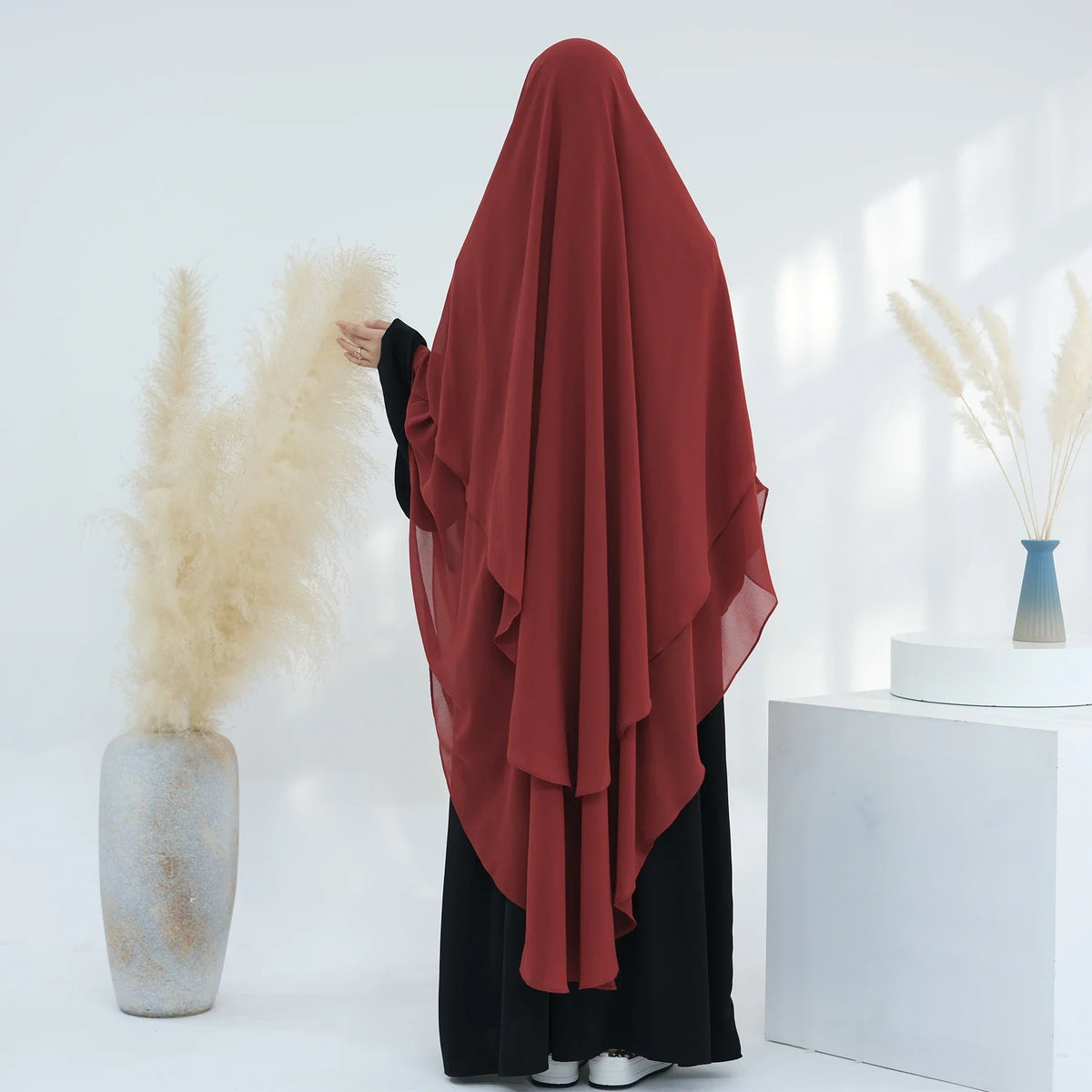 Eine Frau trägt einen tiefroten, zweilagigen Chiffon-Niqab und schwarze Kleidung neben getrocknetem Ziergras.