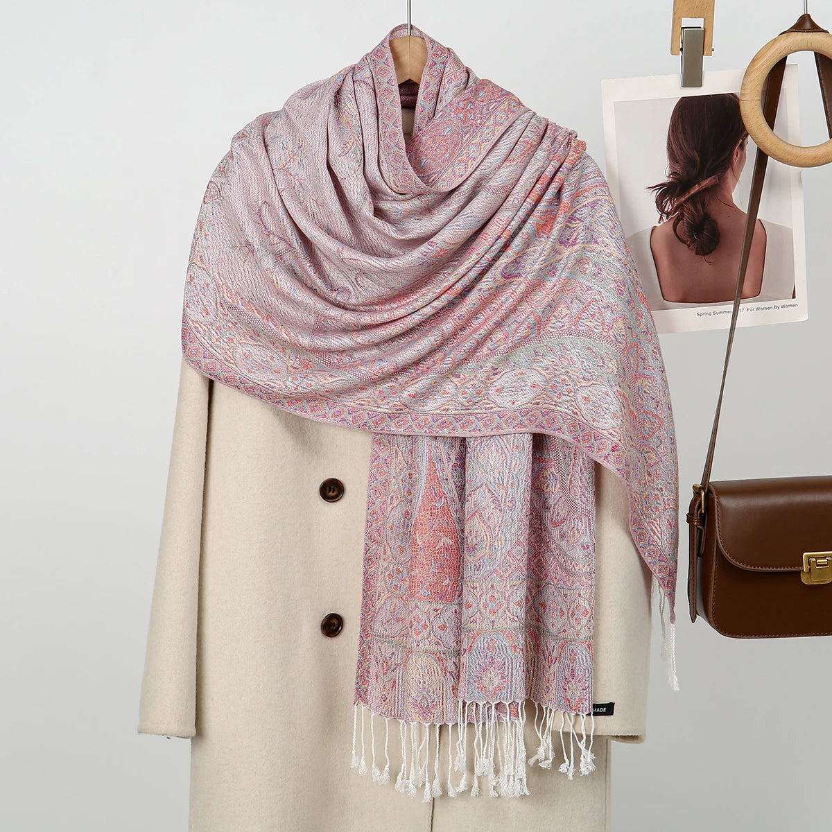 Chal de bordado paisley rosa y coral sobre un abrigo beige, con percha a juego visible.