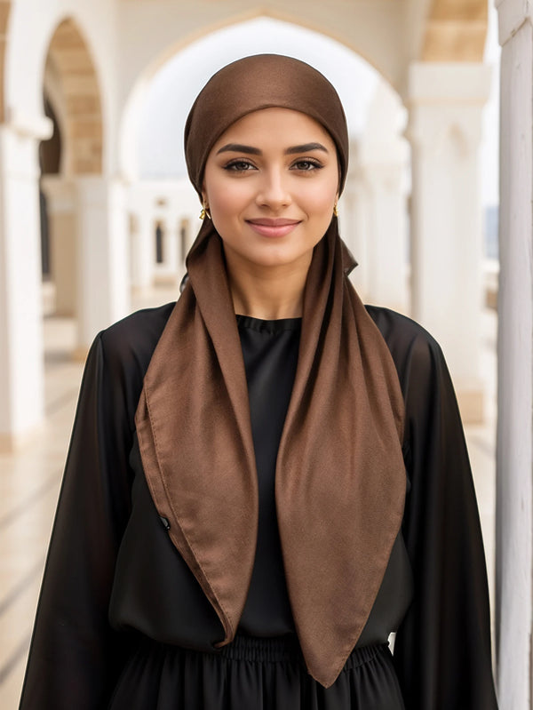 A woman wearing a brown linen inner hijab cap and black clothing stands in an arched outdoor setting.