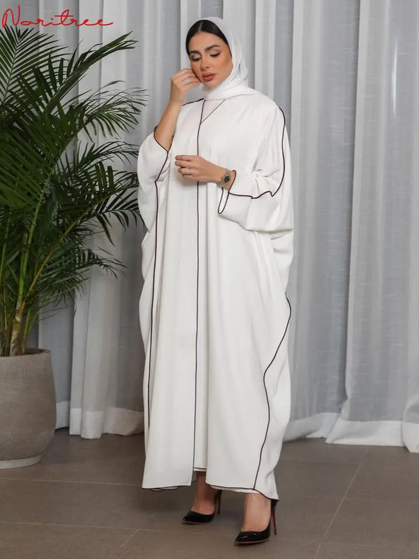 A woman wearing a white, full-length abaya with black trim and a hijab stands indoors.