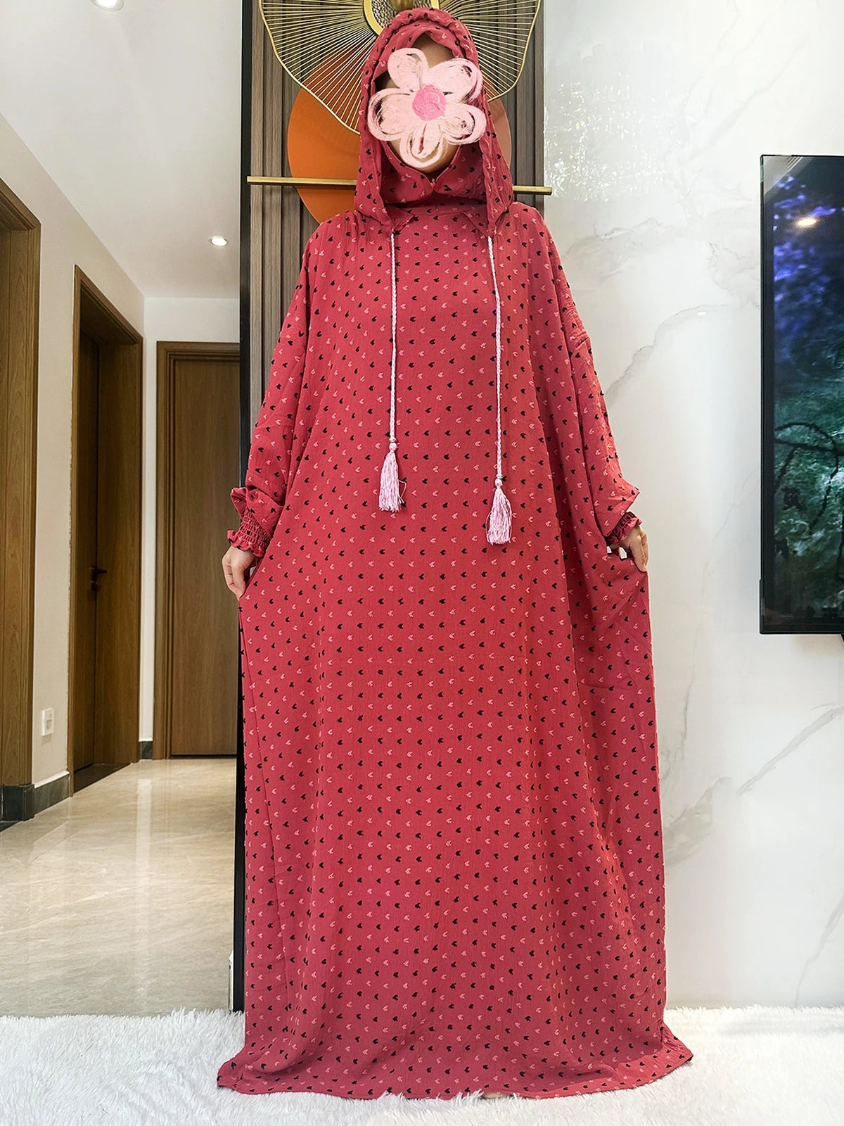 Una mujer vistiendo una abaya de algodón color granate larga con capucha y adornos florales, de pie en un interior luminoso.