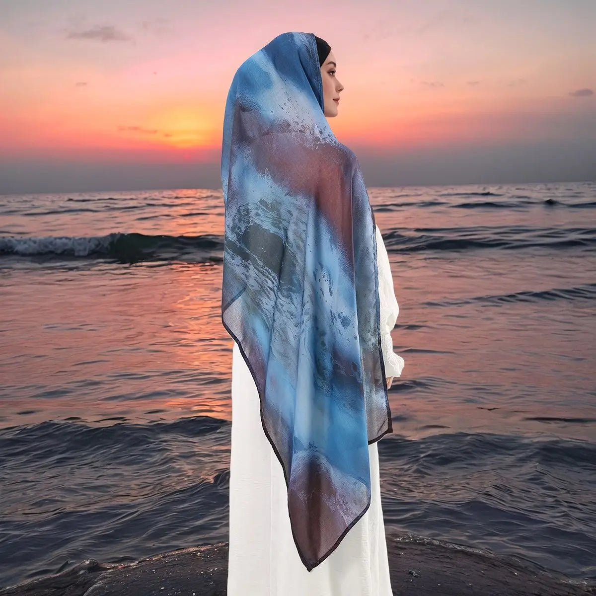 Eine Frau mit blauem Aquarell-digitalem Hijab-Schal steht bei Sonnenuntergang am Meer.
