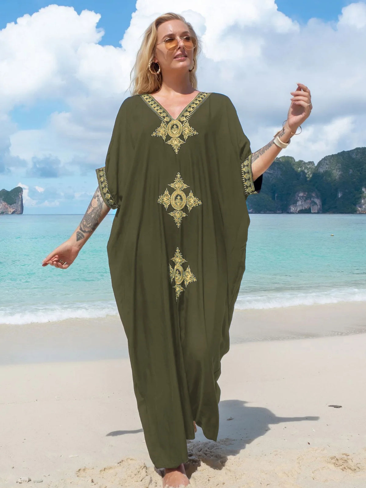 Eine Frau in einem lockeren olivgrünen Kaftan mit Goldstickerei geht am Sandstrand nahe tropischer Inseln.