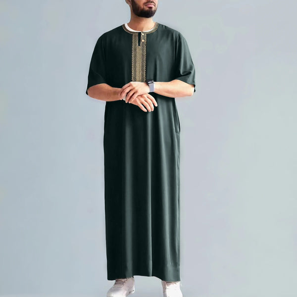A man wearing a dark green embroidered jubbah thobe with a light collar and white shoes.