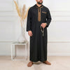 A man wearing a black Moroccan embroidered thobe jellaba with gold detailing, standing against a white wall.
