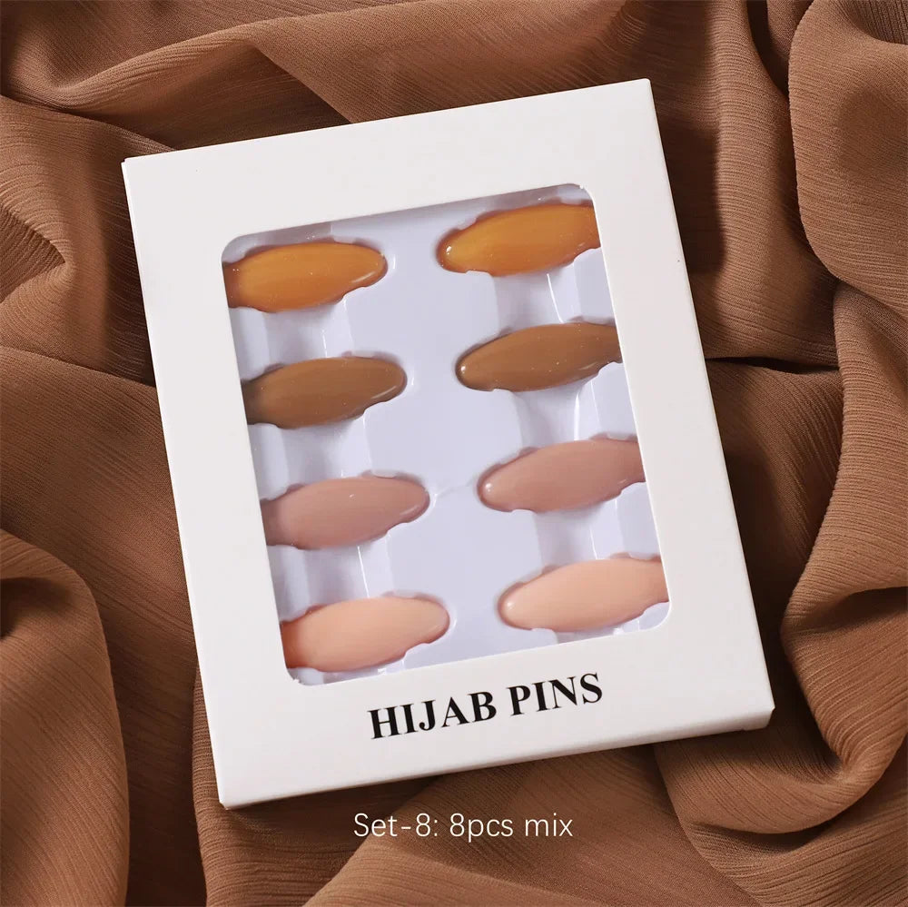Ein Set von acht beigen Hijab-Pins in einer weißen quadratischen Box auf einem braunen Stoffhintergrund.