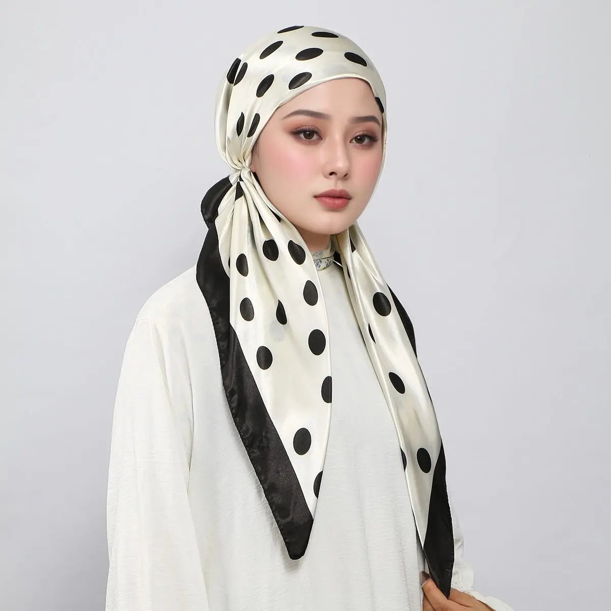 Eine Frau trägt einen satinbedruckten Instant-Hijab-Schal mit schwarzen Polkadottern, gebunden im Turban-Stil.