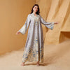 Una mujer modela una abaya larga y fluida con estampado floral gris claro y azul y adornos de diamantes contra un fondo beige