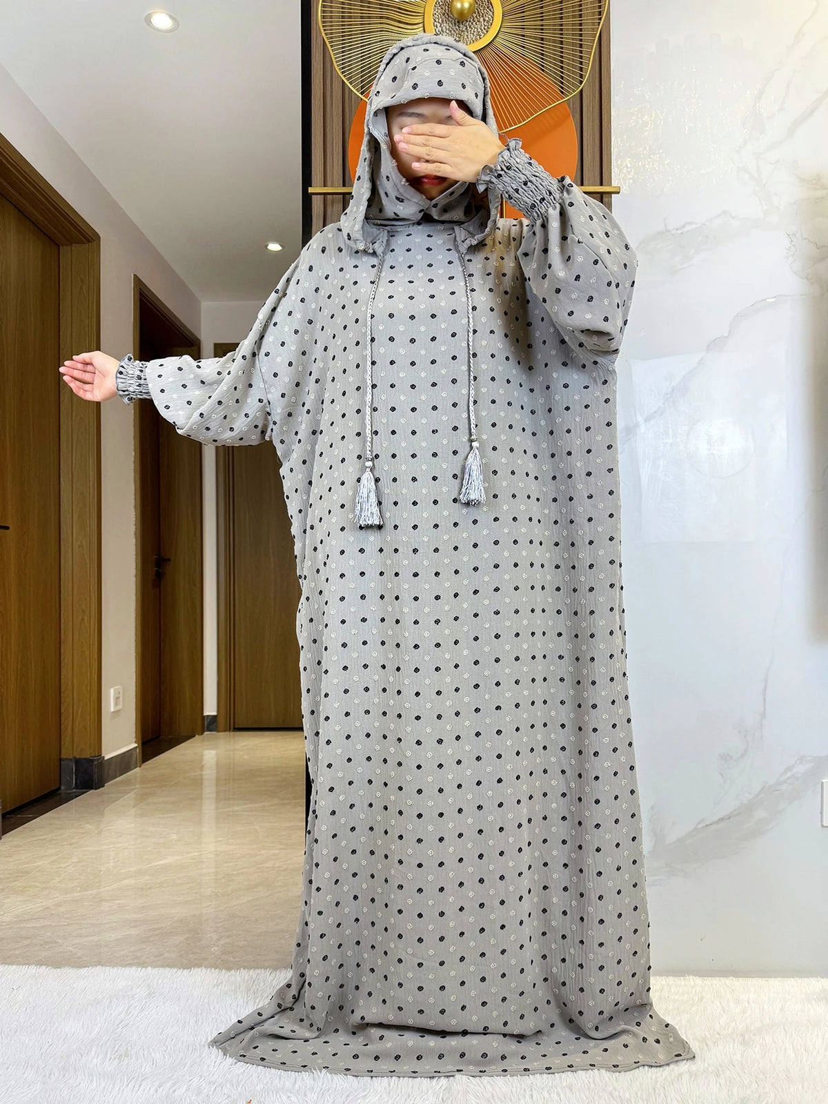Una mujer vistiendo una abaya de algodón gris con puntos, con capucha, de pie en interiores.