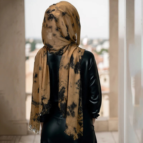 A woman wearing a mustard yellow ombre tie-dye hijab scarf draped over a black top, standing in a bright indoor setting.