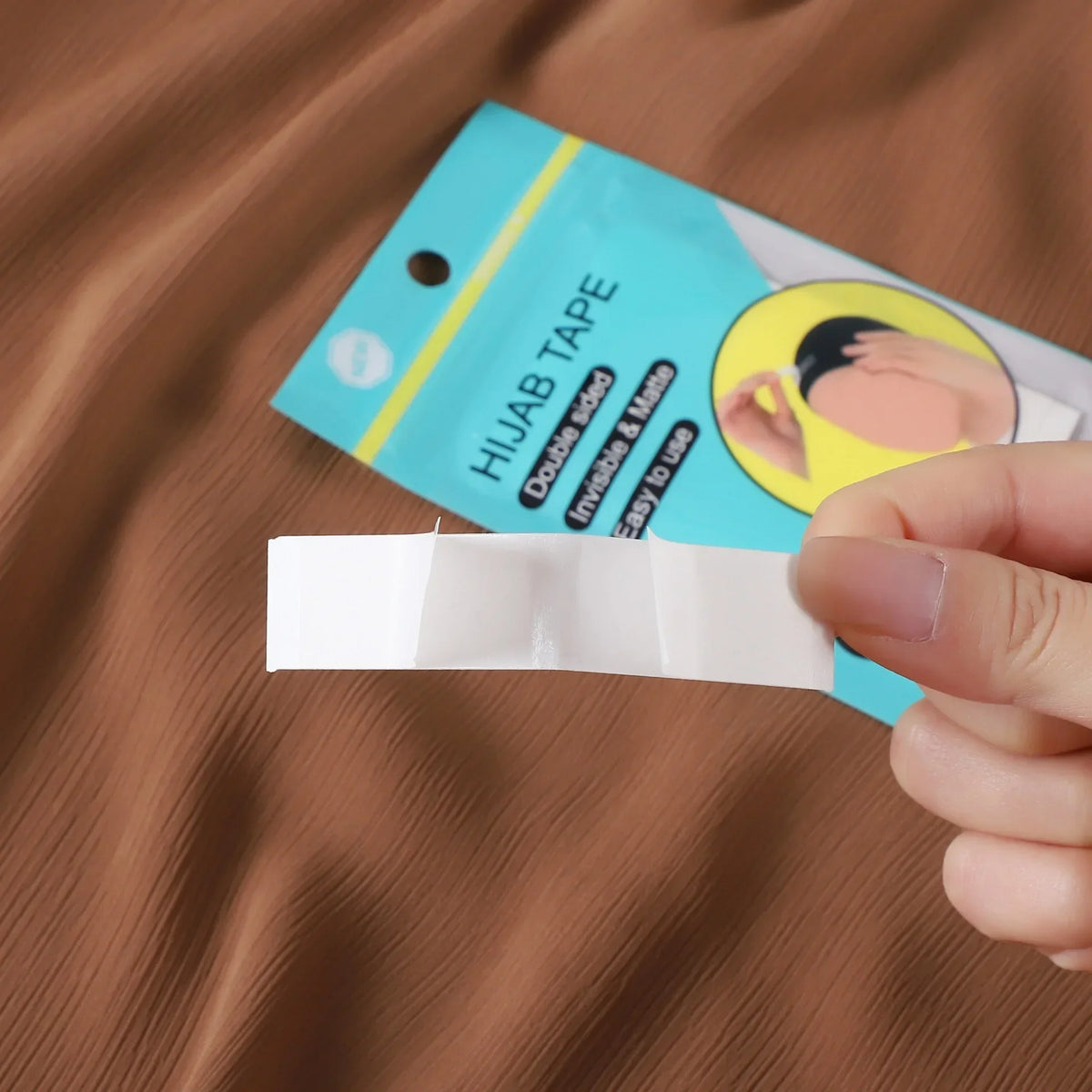 Un rollo de cinta invisible antiadherente para hijab blanca con agarre fuerte, sobre un fondo de tela marrón.