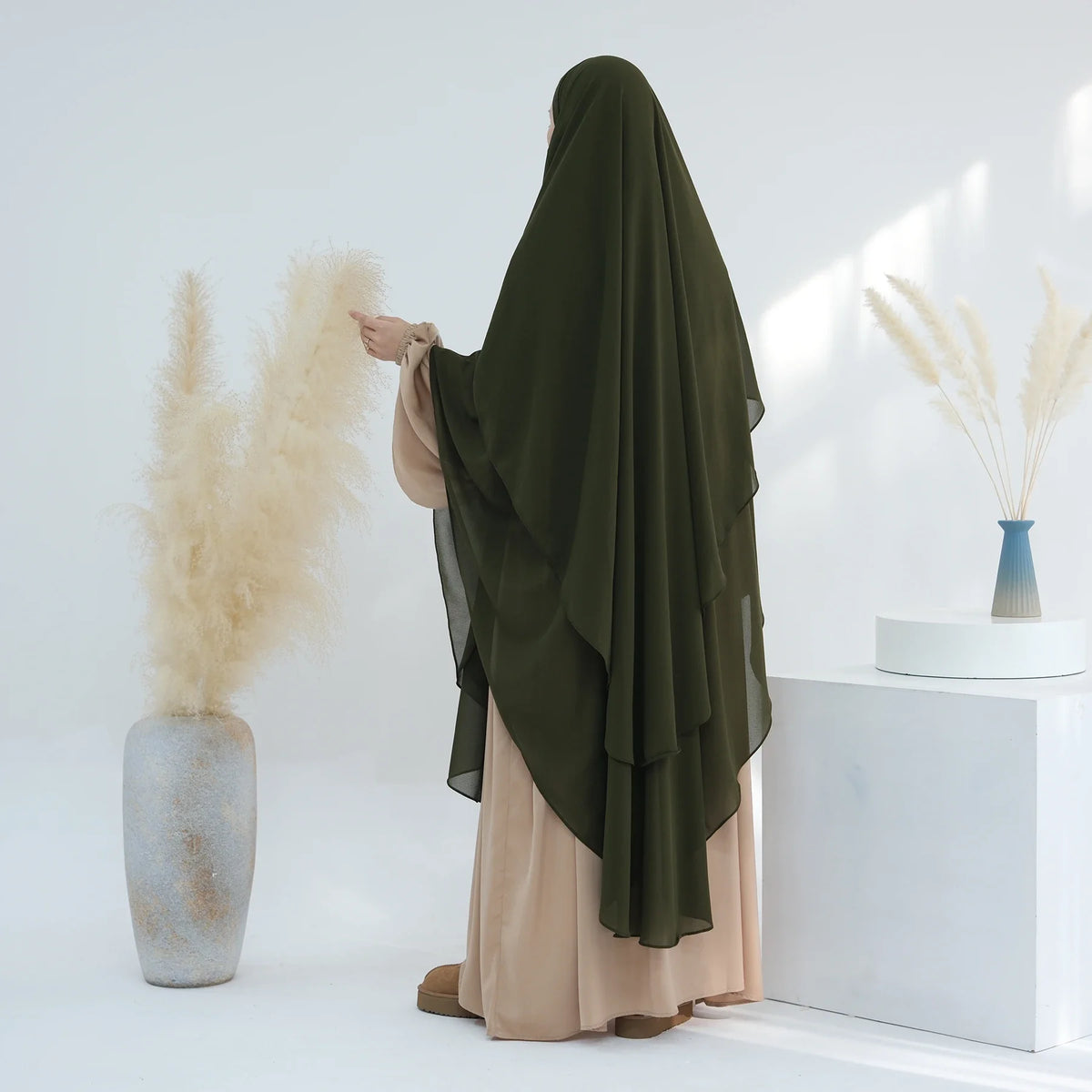 Eine Frau mit dunkel olivgrälichem, zweilagigem Chiffon-Niqab und beige Kleidung steht neben getrocknetem Ziergräsern