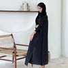 Une femme portant une veste coupe-vent noire plissée se tient à côté d'une étagère blanche et d'une chaise en osier.