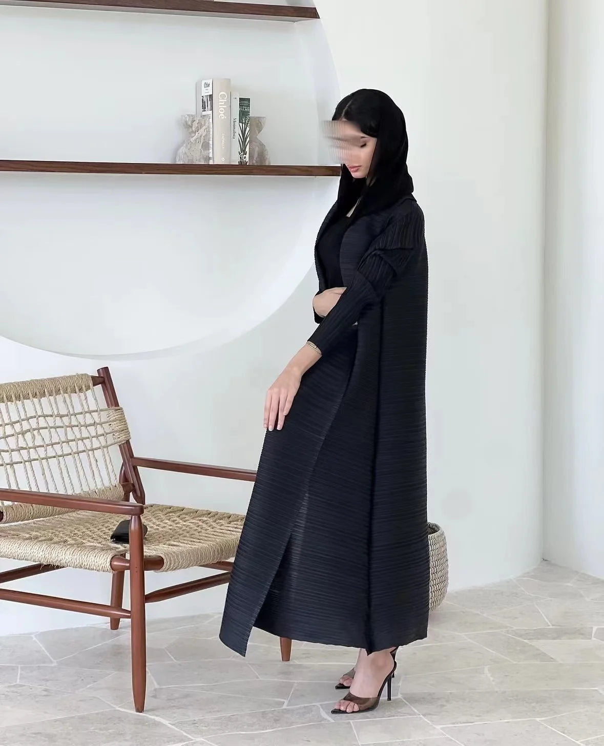Une femme portant une veste coupe-vent noire plissée se tient à côté d'une étagère blanche et d'une chaise en osier.