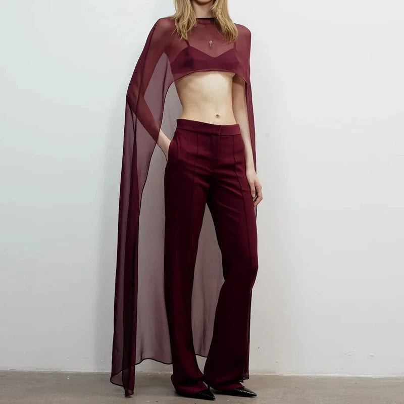 A model wears a deep burgundy sheer mesh poncho cape top and matching trousers against a white background.