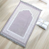 Alfombra de oración rectangular lavanda con patrones geométricos blancos y flecos, junto a un paño blanco doblado.
