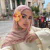 Une femme portant un hijab en mousseline rose pâle à bord de dentelle, regardant directement l'appareil photo.