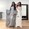 Dos mujeres modelan un abaya de satén plateado con corte batwing y una prenda blanca fluida en una habitación brillante.