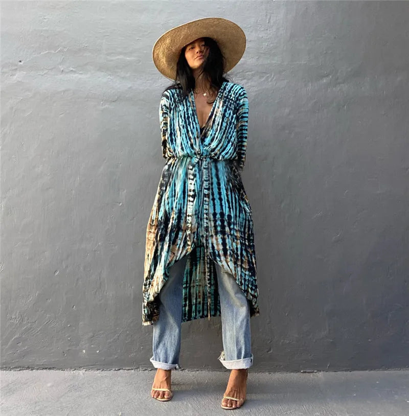 Eine Frau in blau-schwarzem Tie-Dye Kimono-Überkleid und breiter Strohhut vor einer grauen Wand.
