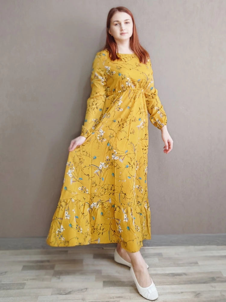 Eine Frau in einem gelben, bohemienartigen Blumenmuster-Maxikleid vor einer grauen Wand.