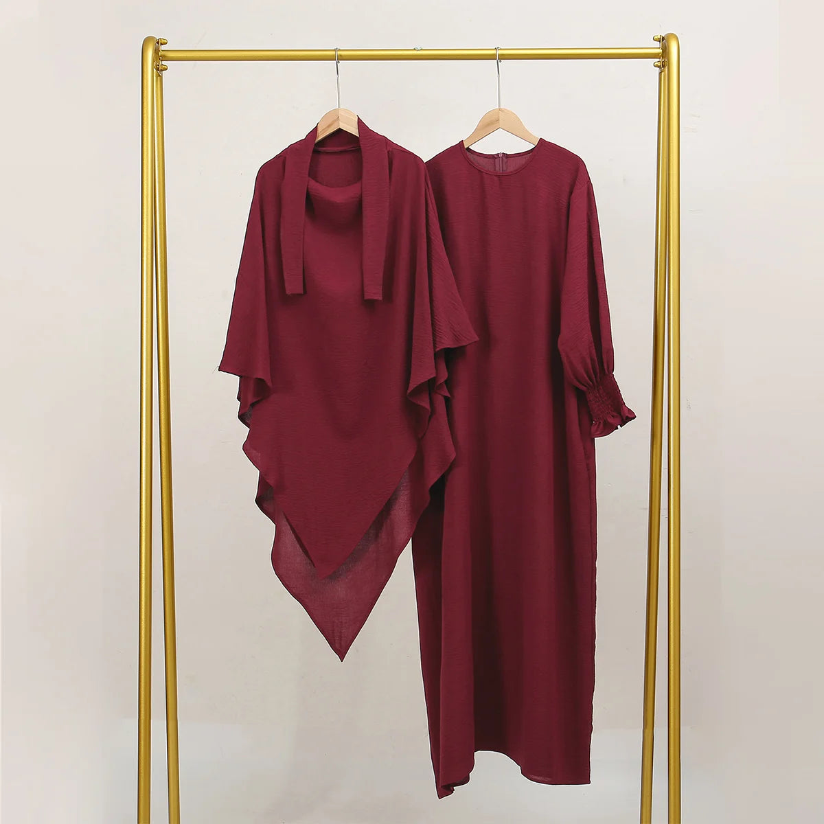 A maroon abaya and khimar set displayed on a gold hanger against a light background.
