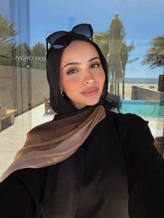 Eine Frau mit schwarzem Hijab und Sonnenbrille posiert vor einer Glaswand mit Palmen und Wasser.