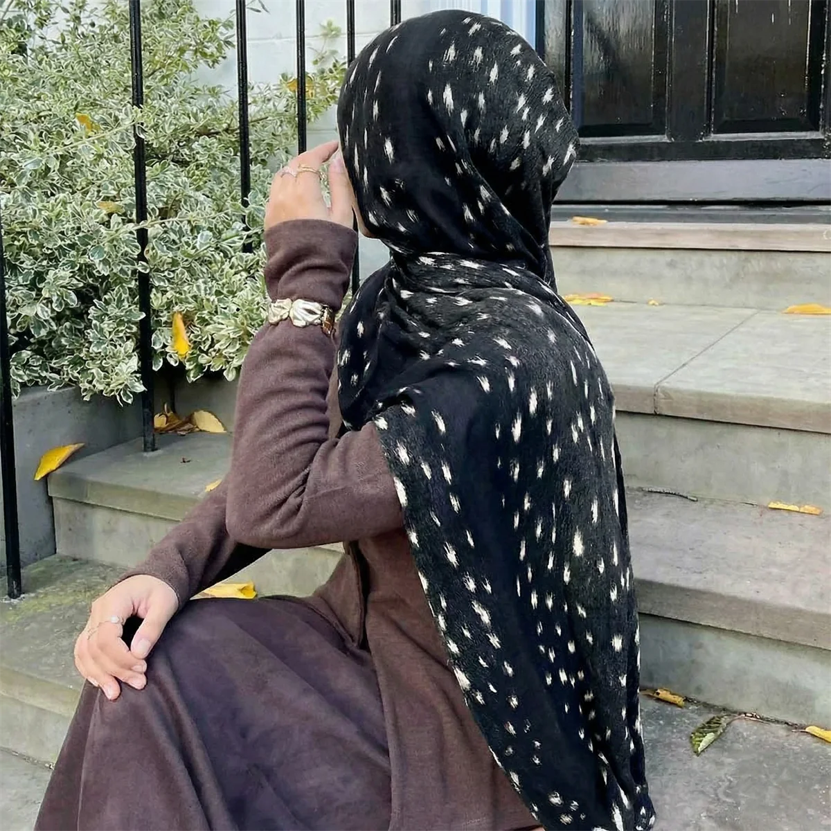 Eine Frau trägt einen schwarzen Hijab mit weißem Hirschmusterdruck und braune Kleidung, sitzt auf Steinstufen.