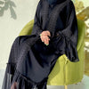 Eine Frau trägt eine elegante schwarze, bodenlange Abaya mit Spitzenverzierung und sitzt in einem grünen Sessel.