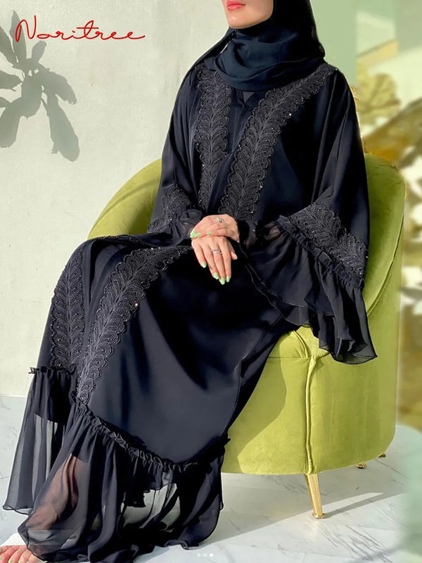 A woman wearing an elegant black full-length abaya with lace detailing, seated in a green armchair.