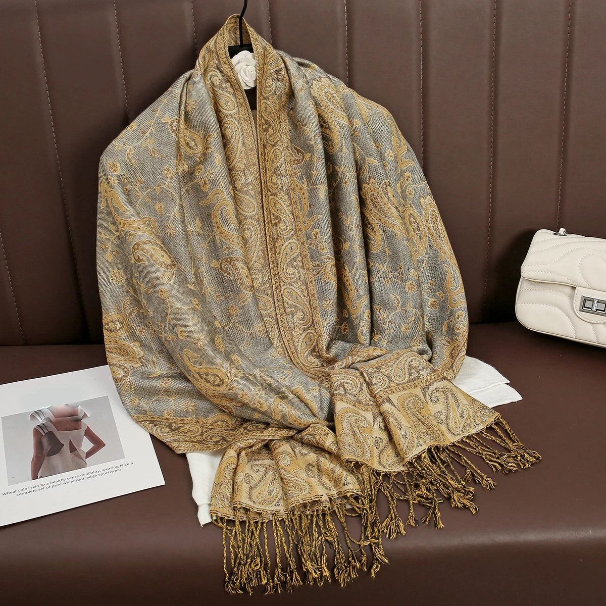 Pañuelo de pashmina lujoso estampado beige y dorado sobre un sofá de cuero marrón.