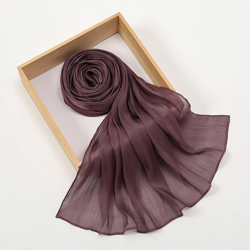 Un chal hijab de gasa plisada color ciruela oscuro enrollado y drapeado en un marco de madera clara.