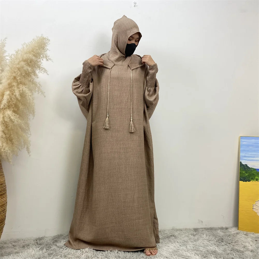 Un mannequin portant une abaya beige avec capuche et manches batwing, debout contre un mur blanc.