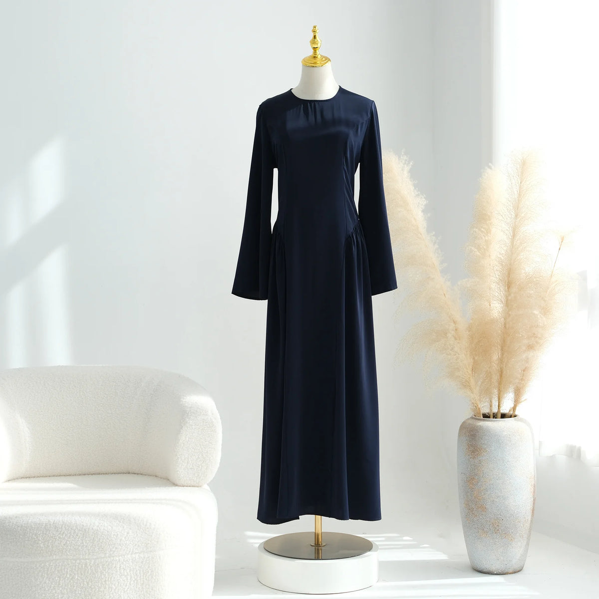 Una elegante abaya de manga larga azul marino, exhibida en un maniquí junto a hierba pampas beige seca y un sofá blanco.