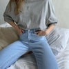 Una persona viste una camiseta básica de algodón gris claro y jeans azules mientras está sentada en una cama.