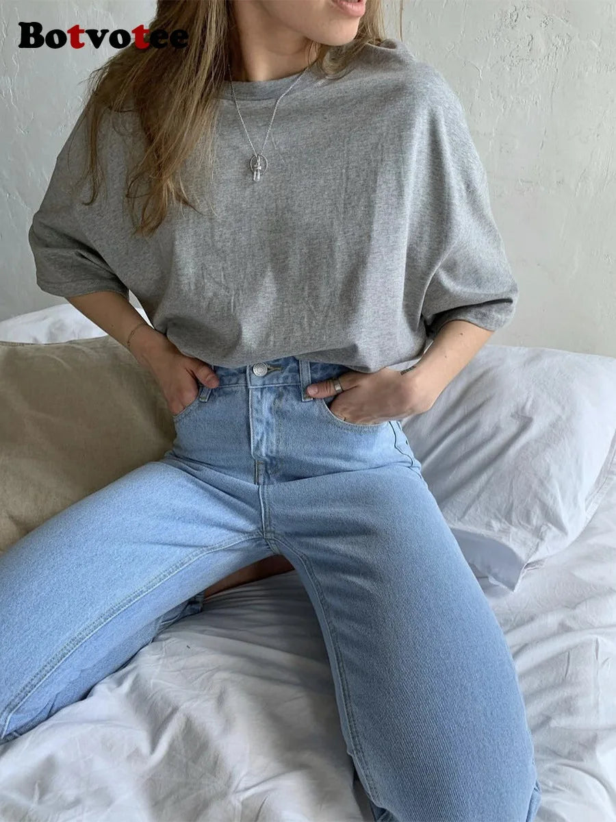 Una persona viste una camiseta básica de algodón gris claro y jeans azules mientras está sentada en una cama.