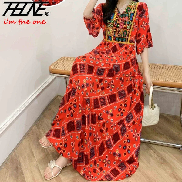 A woman wearing a vibrant orange and red bohemian embroidered maxi dress standing indoors.