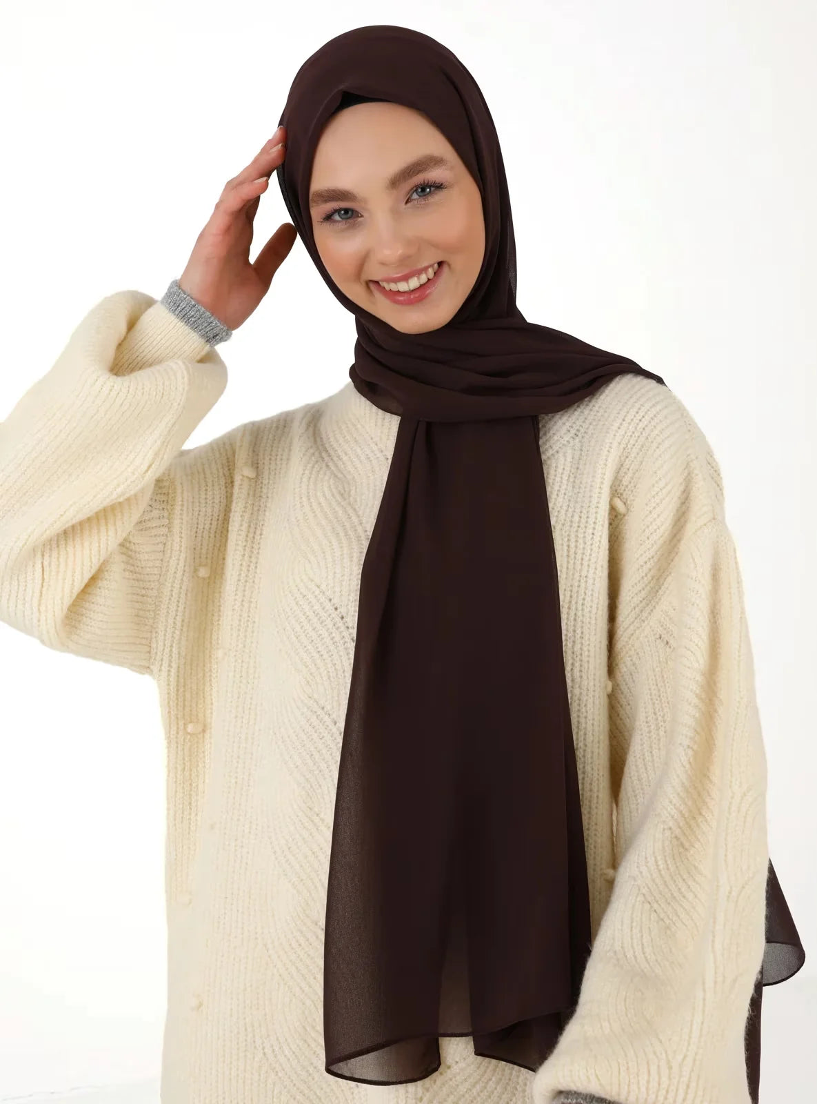 Eine Frau trägt einen dunkelbraunen Chiffon-Hijab und einen cremefarbenen Strickpullover.