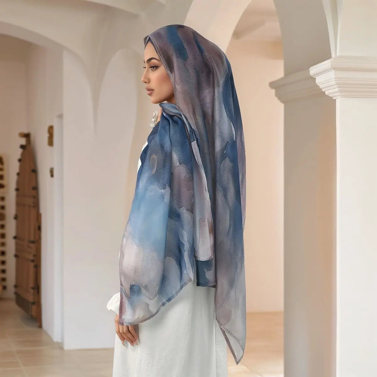 Eine Frau mit einem Aquarell-Digitaldruck-Hijabschal in Blau- und Grautönen steht in einem hellen Flur.