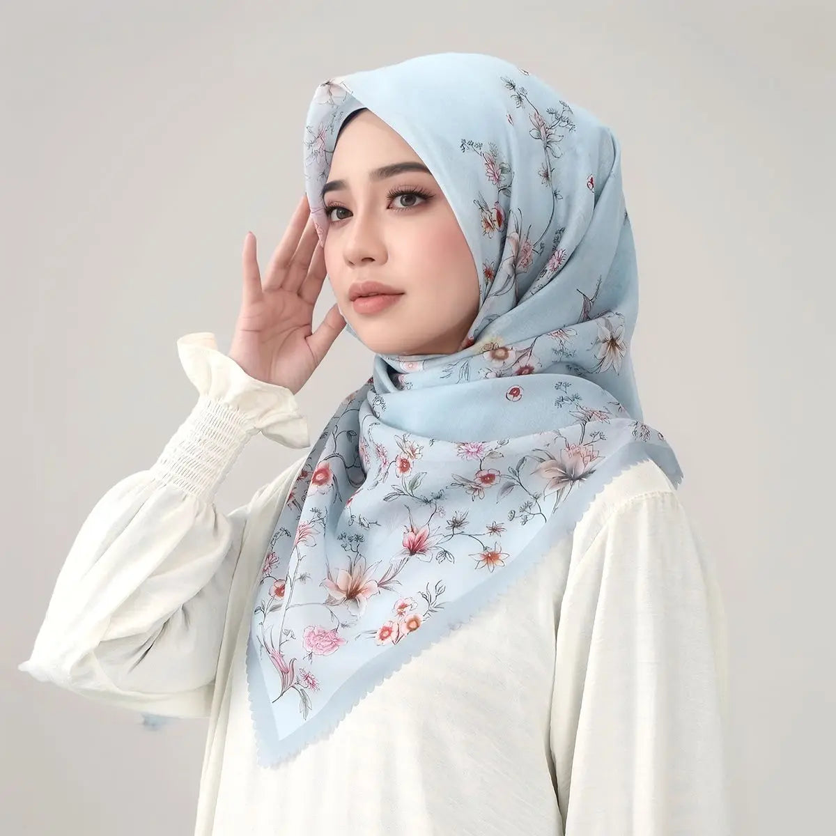 Eine Frau trägt einen hellblauen blumigen quadratischen Hijab und ein weißes Oberteil, posiert mit der Hand nahe am Gesicht.
