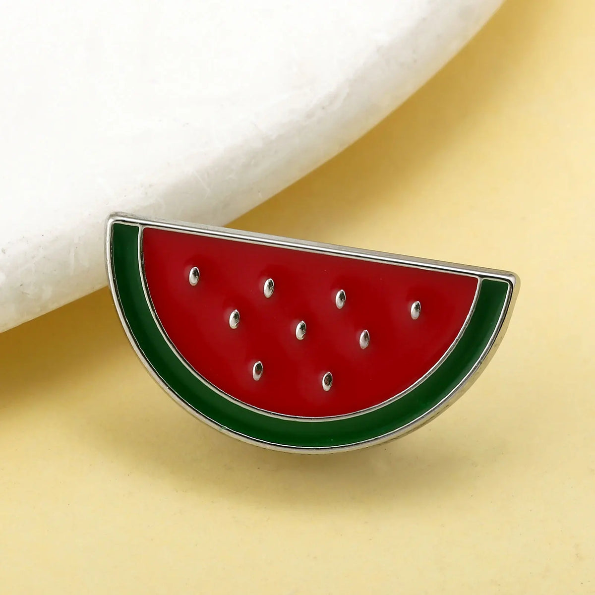 Rotes Wassermelonen-Emaliepenn-Anstecknadel mit grüner Schale und silberner Umrandung auf hellem gelbem Untergrund.