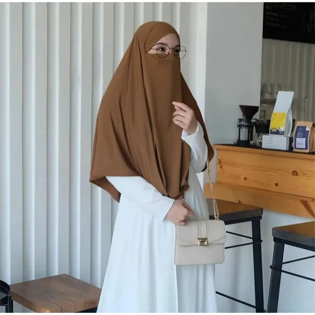 Eine Frau in brachem Hijab und weißem Kleid wird drinnen gezeigt.