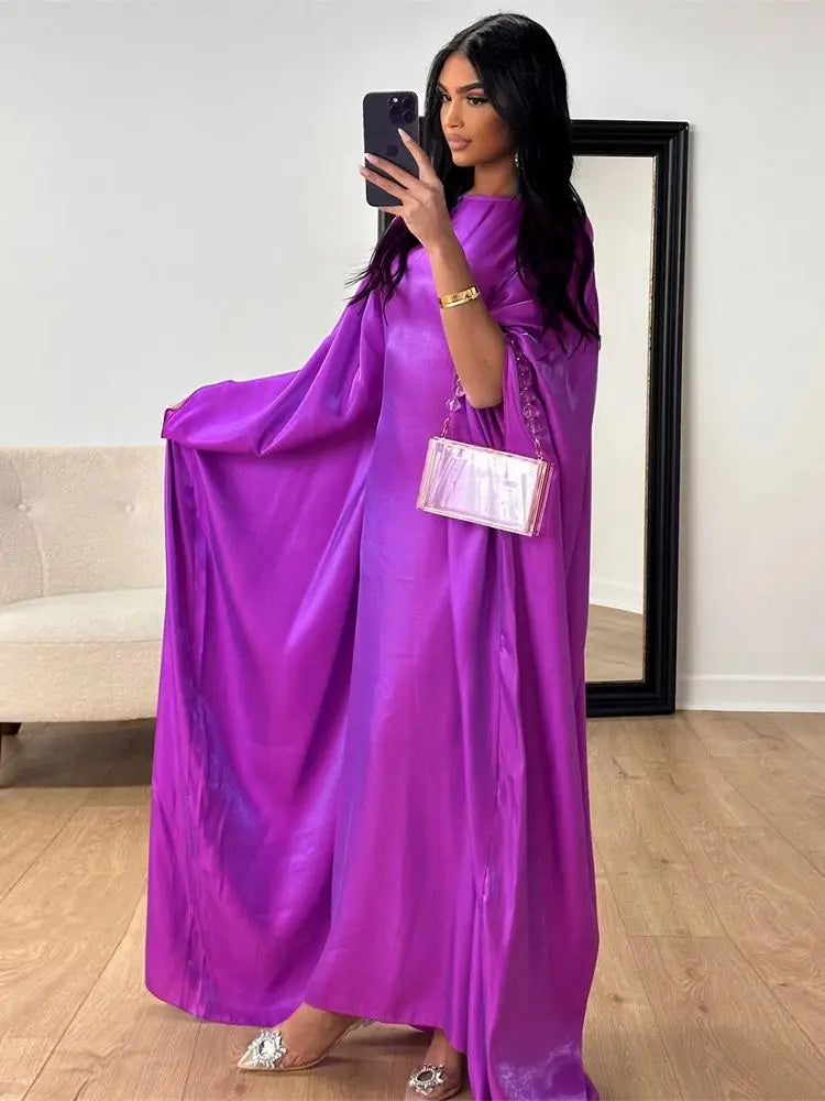 Una mujer modela una vibrante abaya de satén morado batwing con una pulsera dorada y un bolso transparente.
