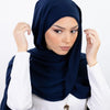 Une femme portant un hijab en mousseline bleu marine et un haut blanc, regardant directement l'appareil photo.