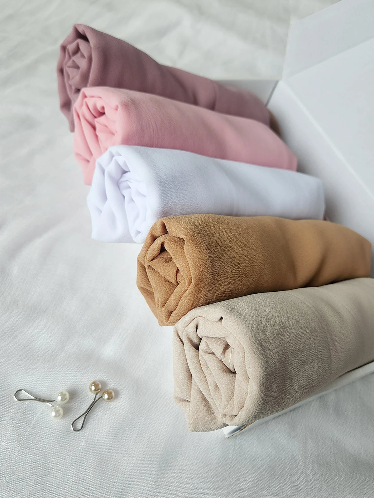 Un juego de pañuelos de chifón doblados en tonos rosa, beige y blanco, dispuestos dentro de una caja blanca.