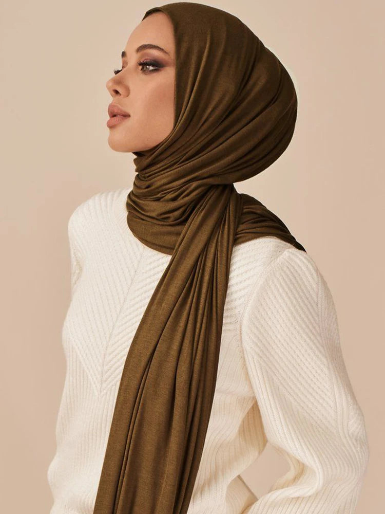 Eine Frau trägt einen olivgrünen Modal-Jersey-Hijabschal aus Baumwolle und einen weißen Strickpullover.